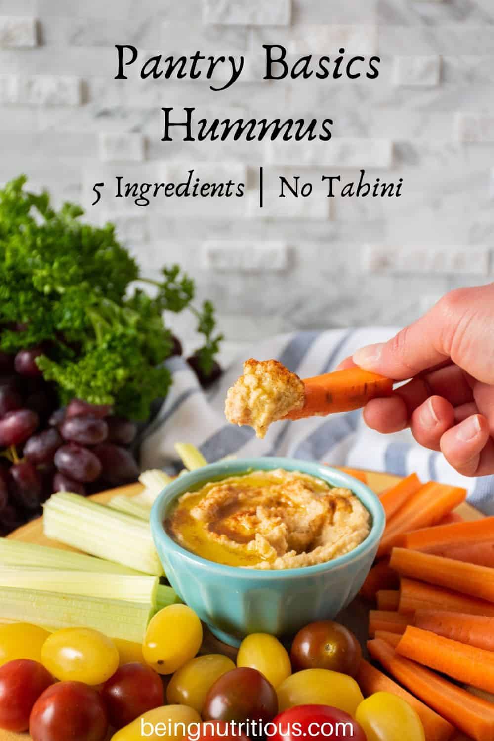 Bowl of pantry basics hummus in the middle of a vegetable platter, with a hand dipping a carrot stick in the hummus.