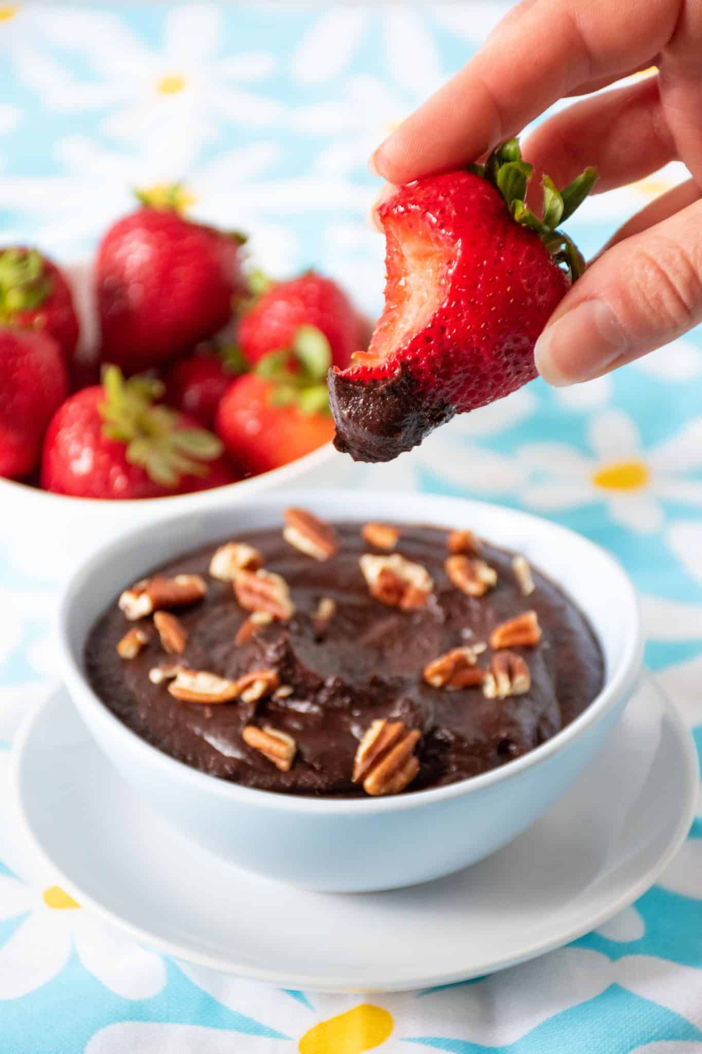 A strawberry with brownie batter dip on it and a bite taken out. Bowl of dip behind.