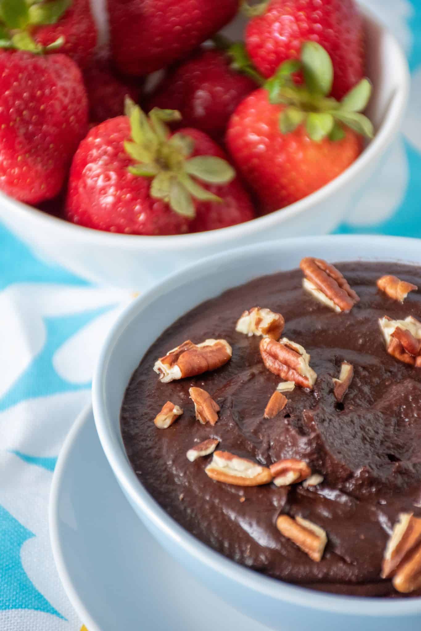 Close up of brownie batter in a bowl with pecan pieces on top. A bowl of strawberries is seen behind.