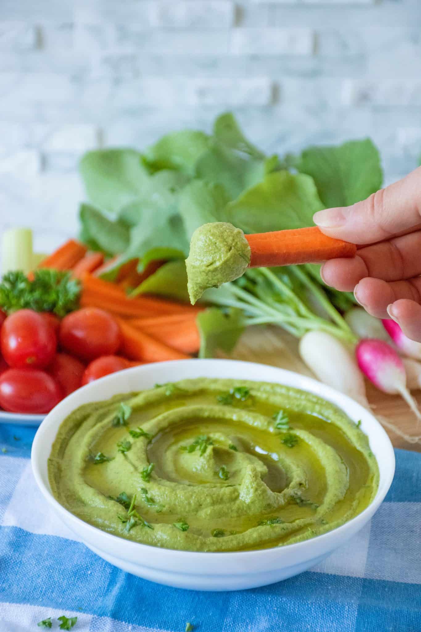 Radish Greens Hummus in a bowl with a hand dipping a carrot stick in it.