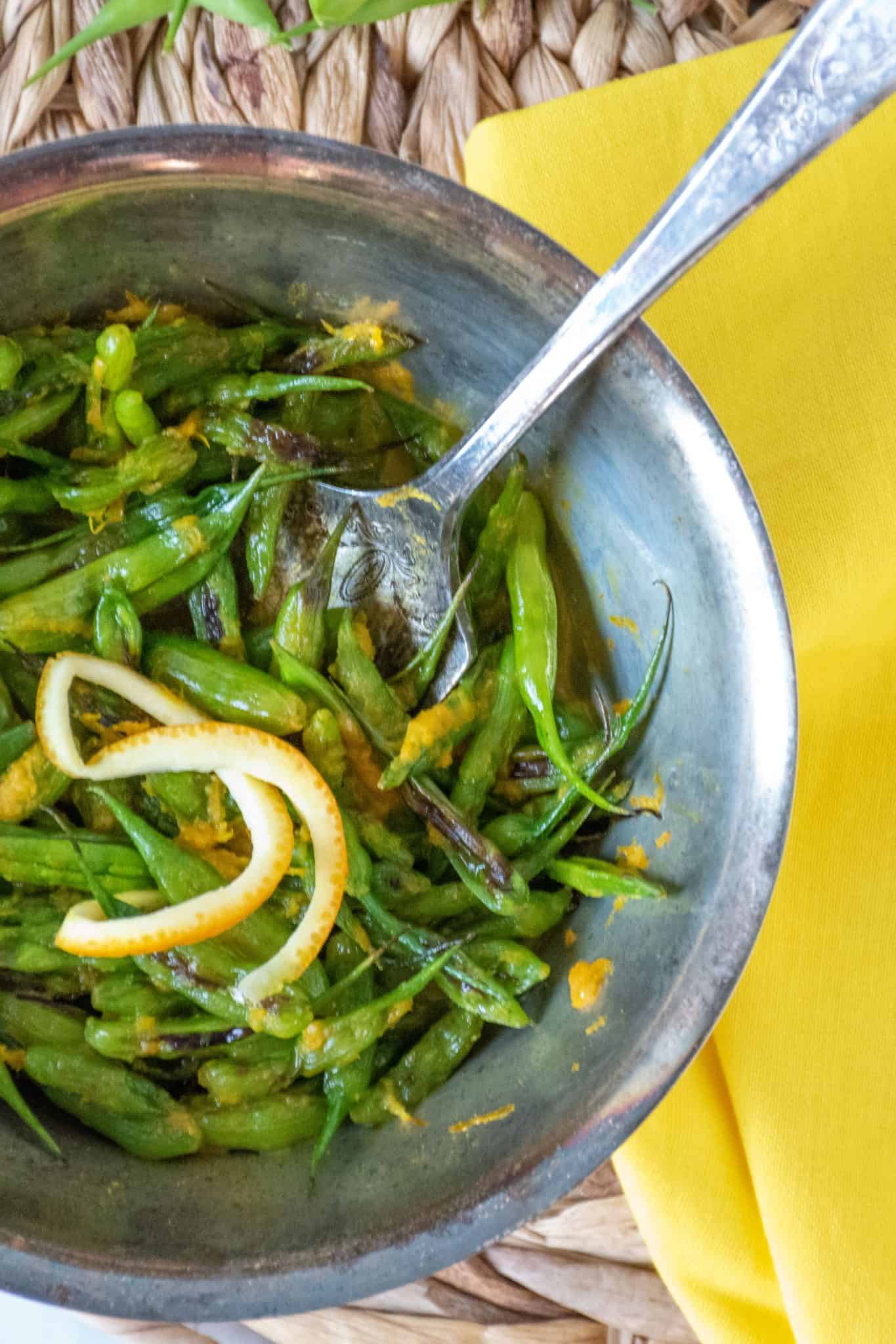 Overhead shot of charred radish pods and orange peel garnish in a silver bowl with a silver fork.