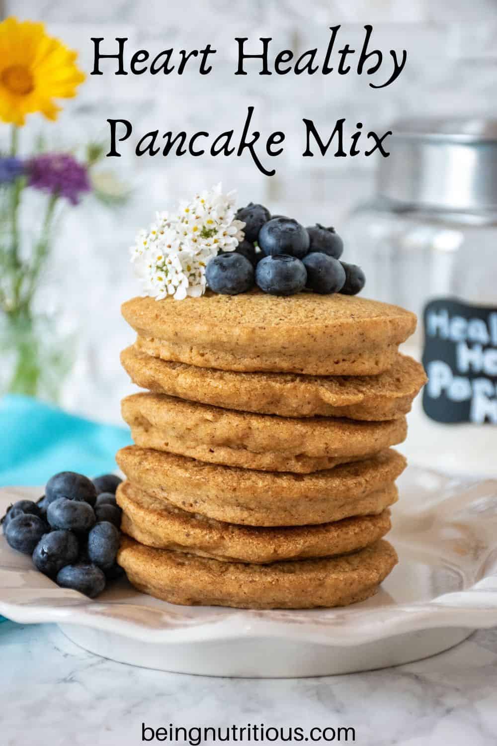 Stack of 6 whole wheat pancakes, with a pile of fresh blueberries and white flowers on top.