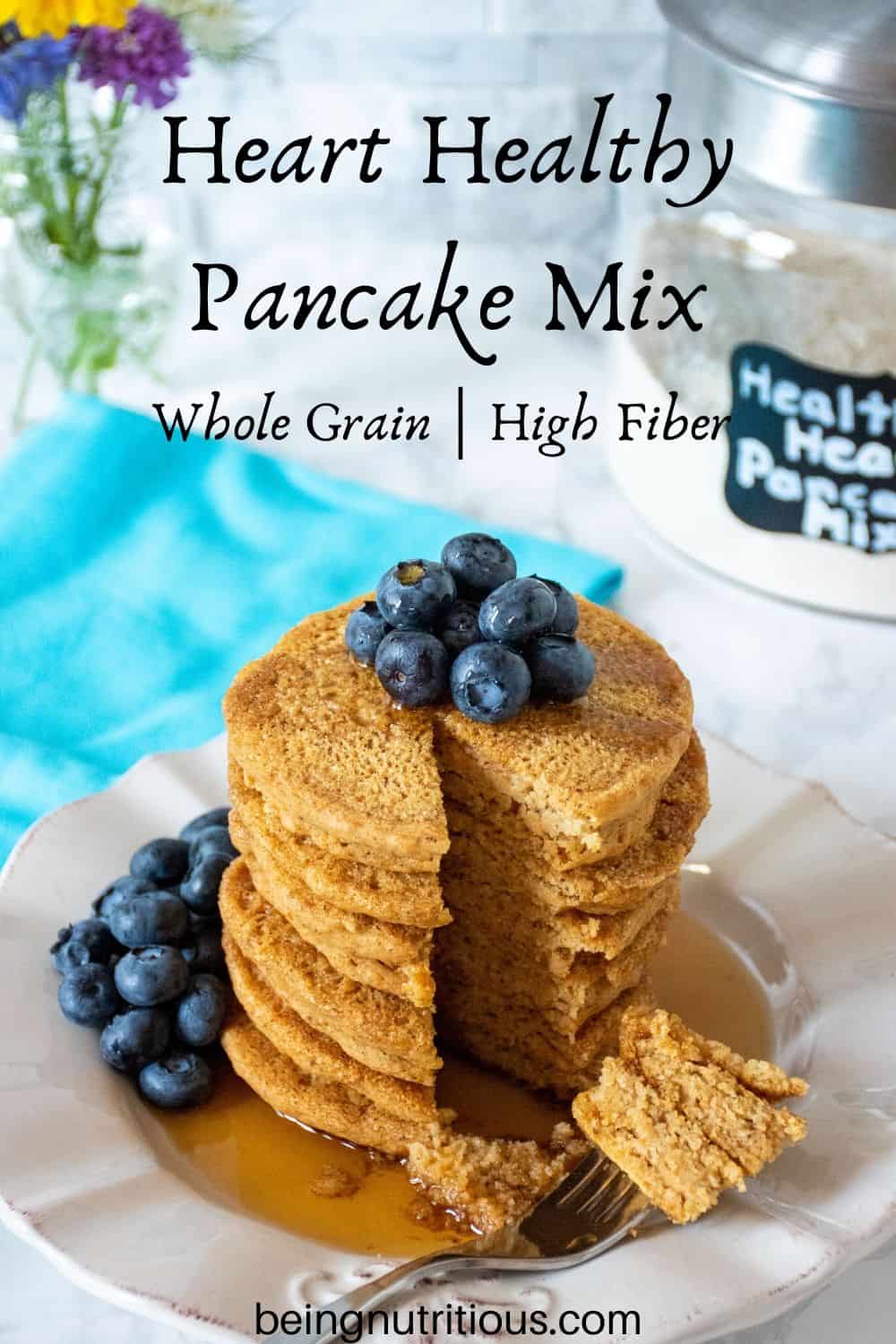 Stack of 6 whole wheat pancakes, with a pile of fresh blueberries on top, and syrup poured over. A fork with cut pieces of pancakes on it lies in front of the stack. Glass jar of heart healthy pancake mix is visible in the background with a bouquet of flowers.