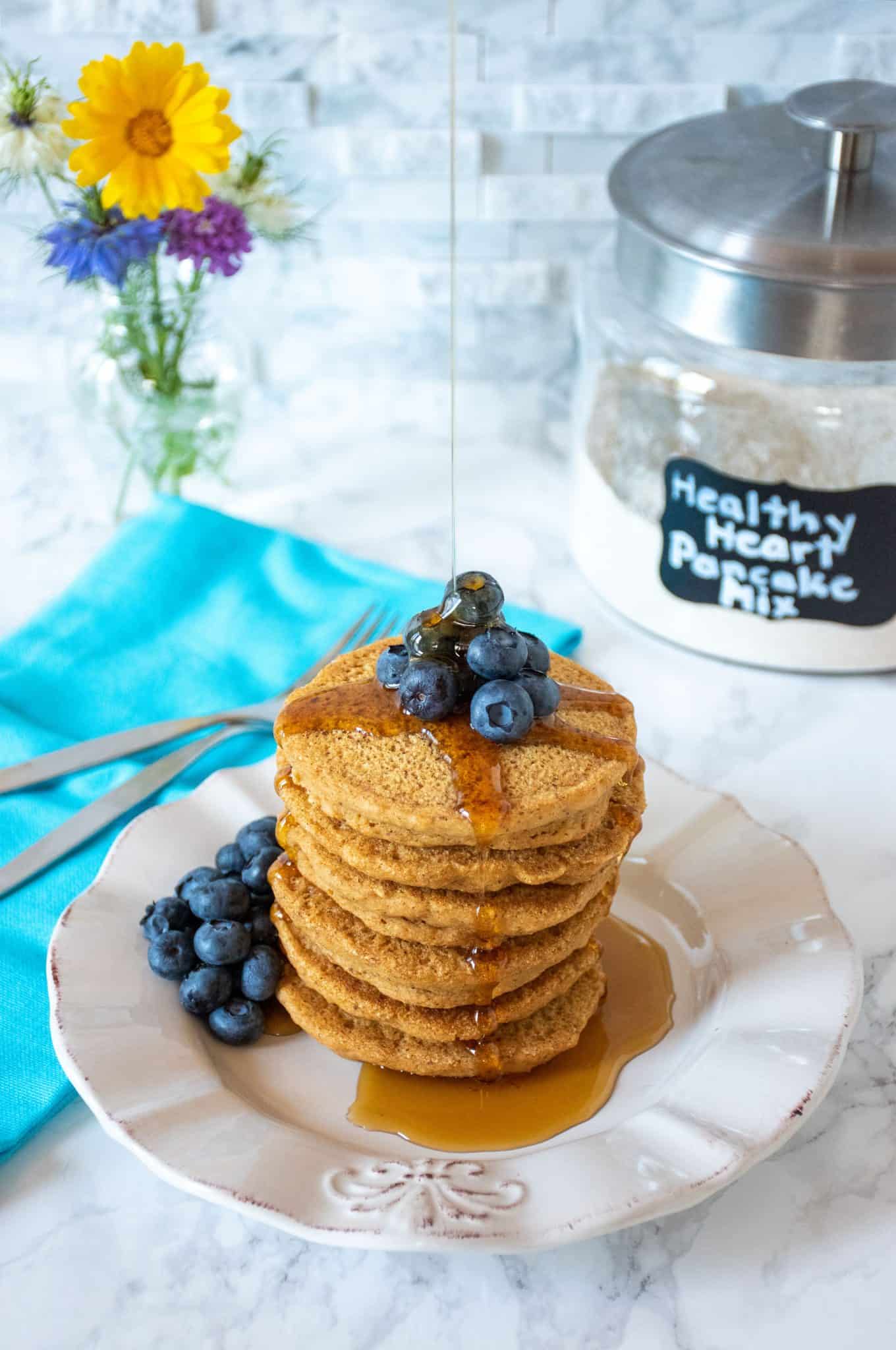 Stack of 6 whole wheat pancakes, with a pile of fresh blueberries on top, and syrup being poured over. Glass jar that reads 'healthy heart pancake mix' is visible in the background.