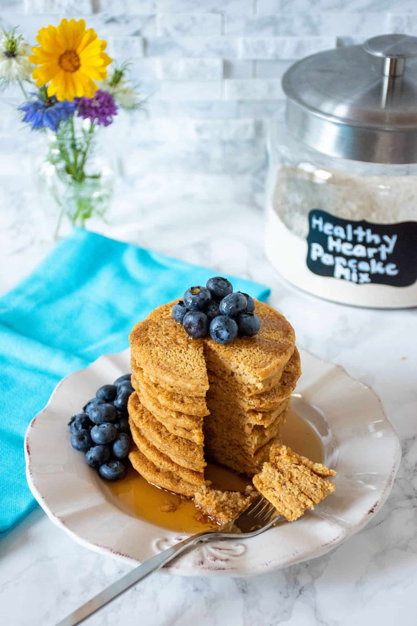 Stack of 6 whole wheat pancakes, with a pile of fresh blueberries on top, and syrup poured over. A fork with cut pieces of pancakes on it lies in front of the stack. Glass jar of heart healthy pancake mix is visible in the background with a bouquet of flowers.