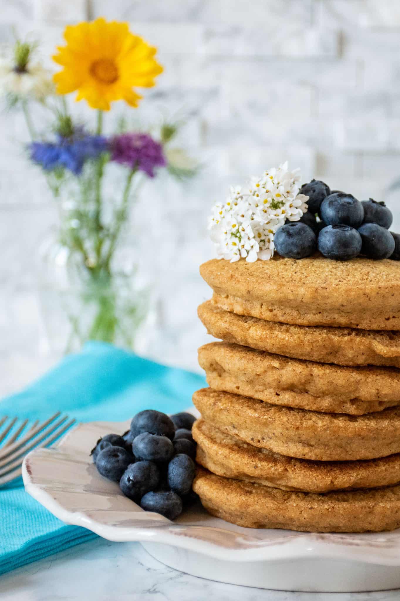 Stack of 6 whole wheat pancakes, with a pile of fresh blueberries and white flowers on top. A small bouquet of wildflowers is visible in the background.