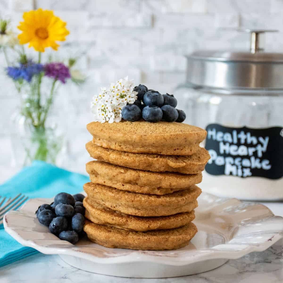 Stack of 6 whole wheat pancakes, with a pile of fresh blueberries on top, and piled to the right of the stack. Glass jar that reads 'healthy heart pancake mix' is visible in the background with a bouquet of wildflowers.