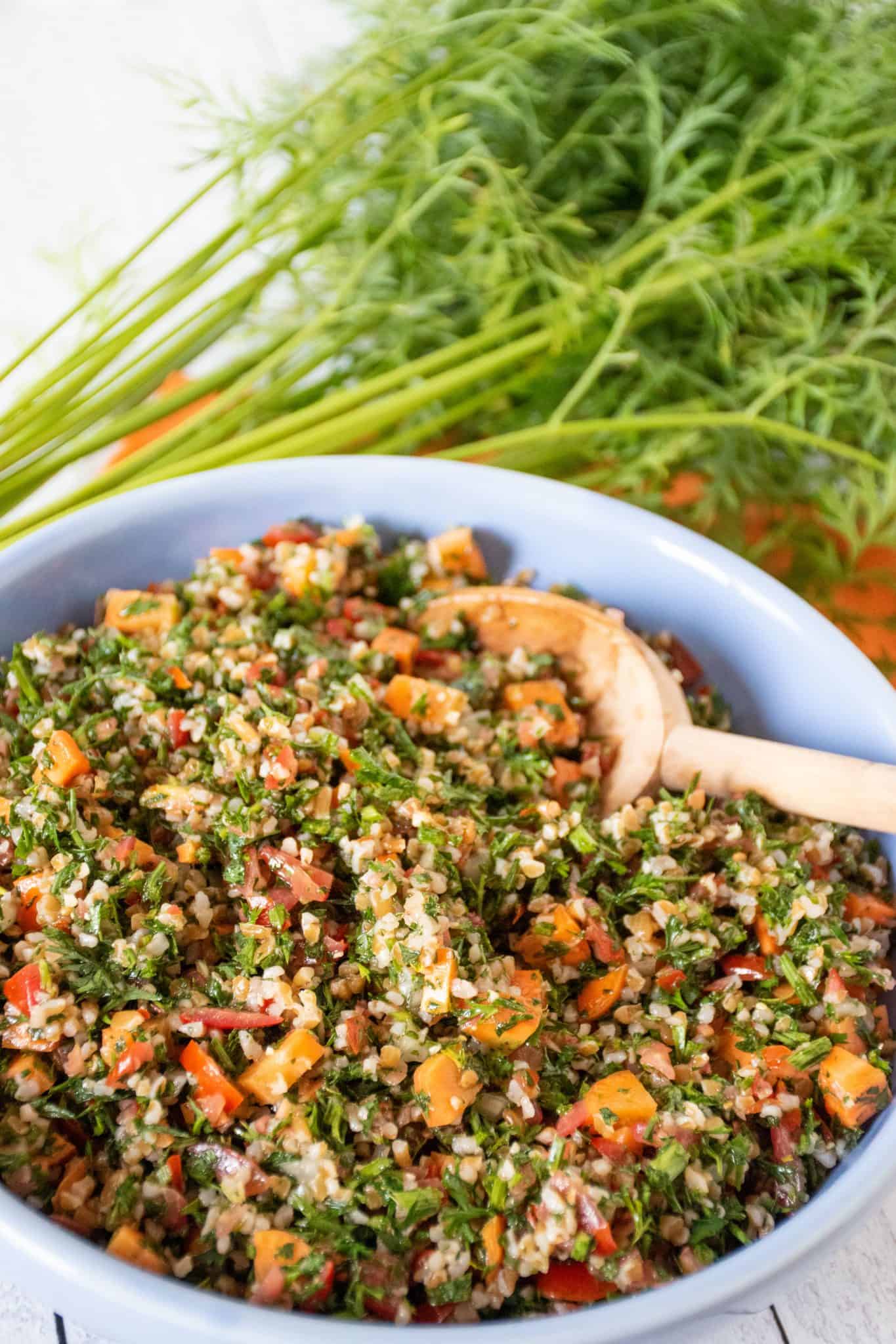 Close up of tabbouleh in blue bowl, with carrot tops visible in background.