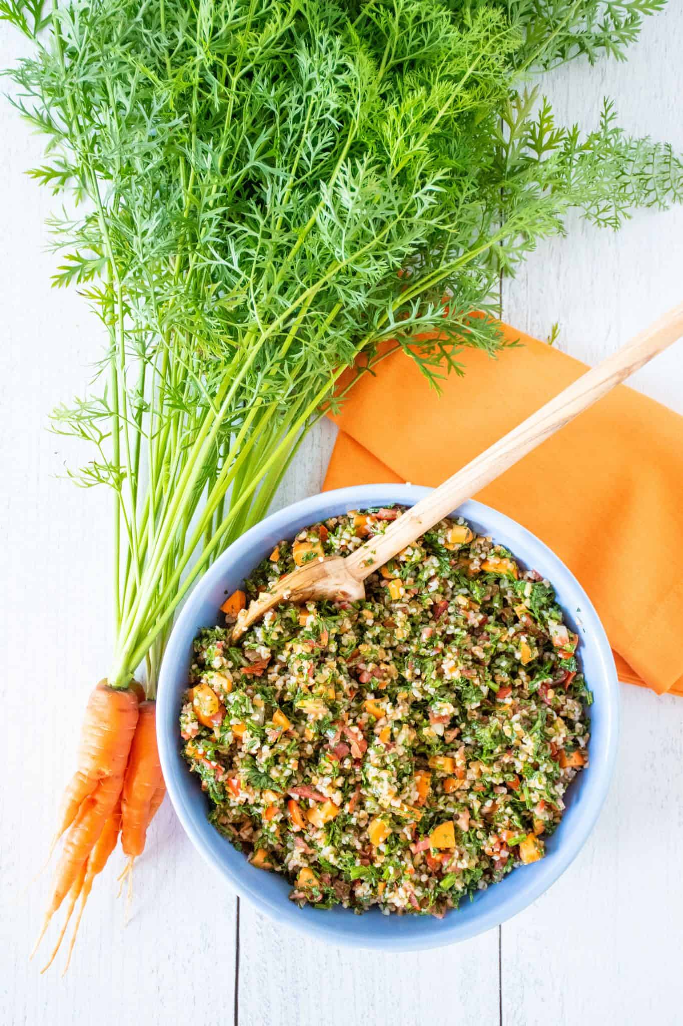 Overhead shot of carrot top tabbouleh in a blue bowl, with a bunch of carrots with the tops still attached lying beside the bowl