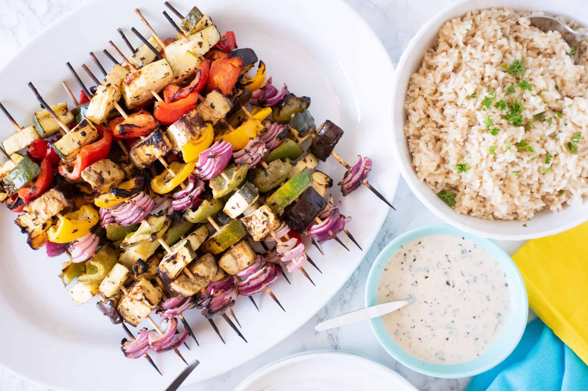 Serving platter with vegetable skewers piled on. A bowl of rice and marinade to the side.