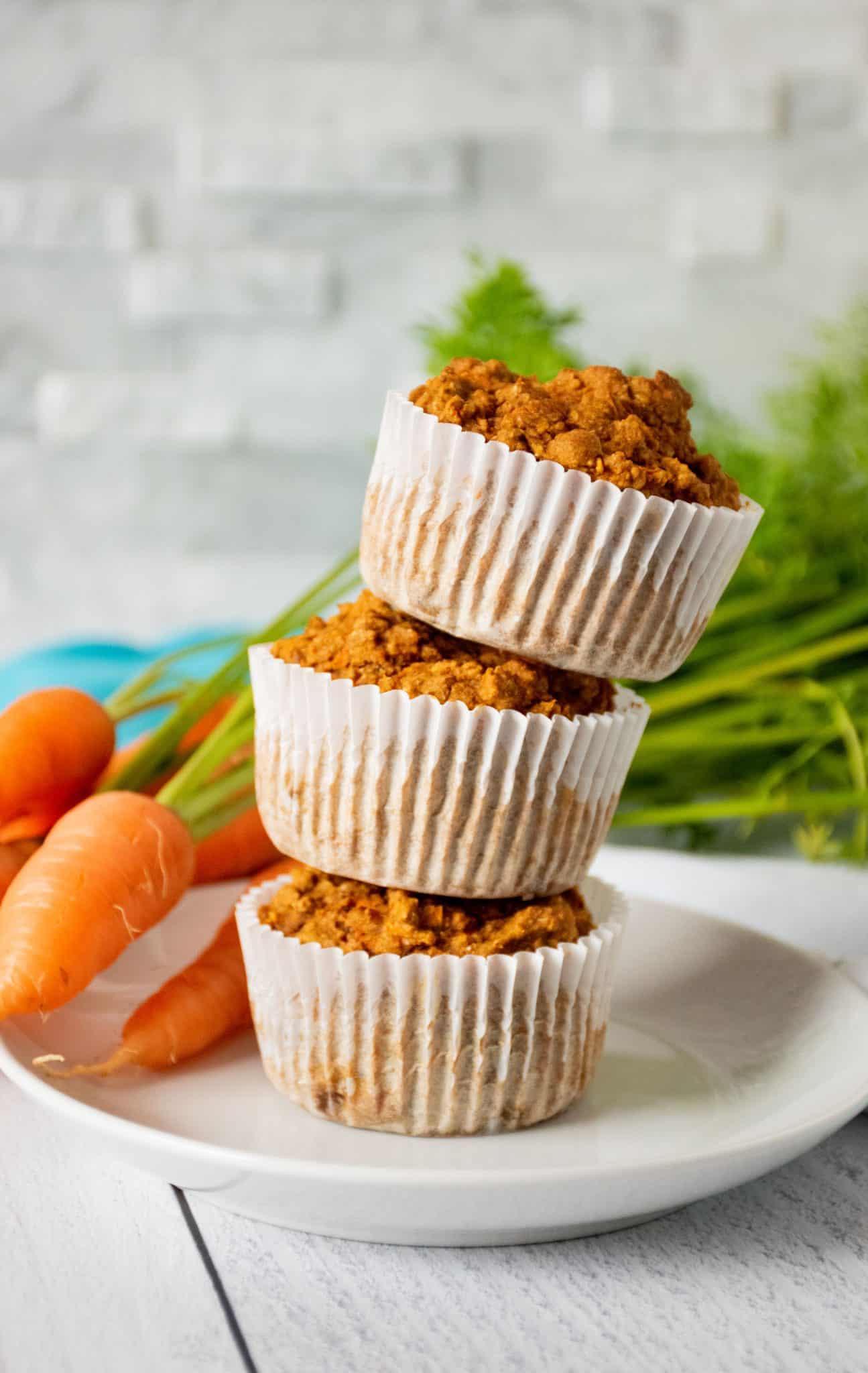 Stack of 3 muffins on a plate with fresh carrots in the background.