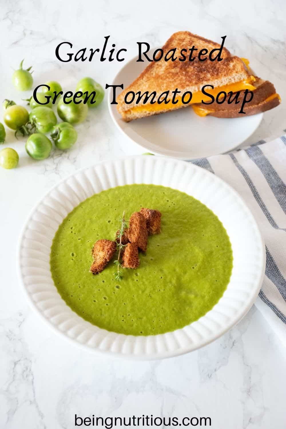 Green soup in a bowl with homemade croutons. Plate with grilled cheese sandwich in background. Text overlay: Garlic Roasted Green Tomato Soup.