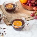 Small glass mugs filled with mulled grape juice, each with a slice of orange in them. Fresh grapes and oranges in background.