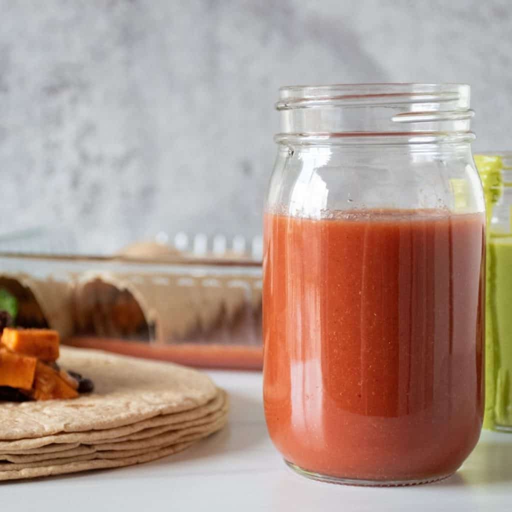 Red sauce in a jar, with enchiladas being made in the background.