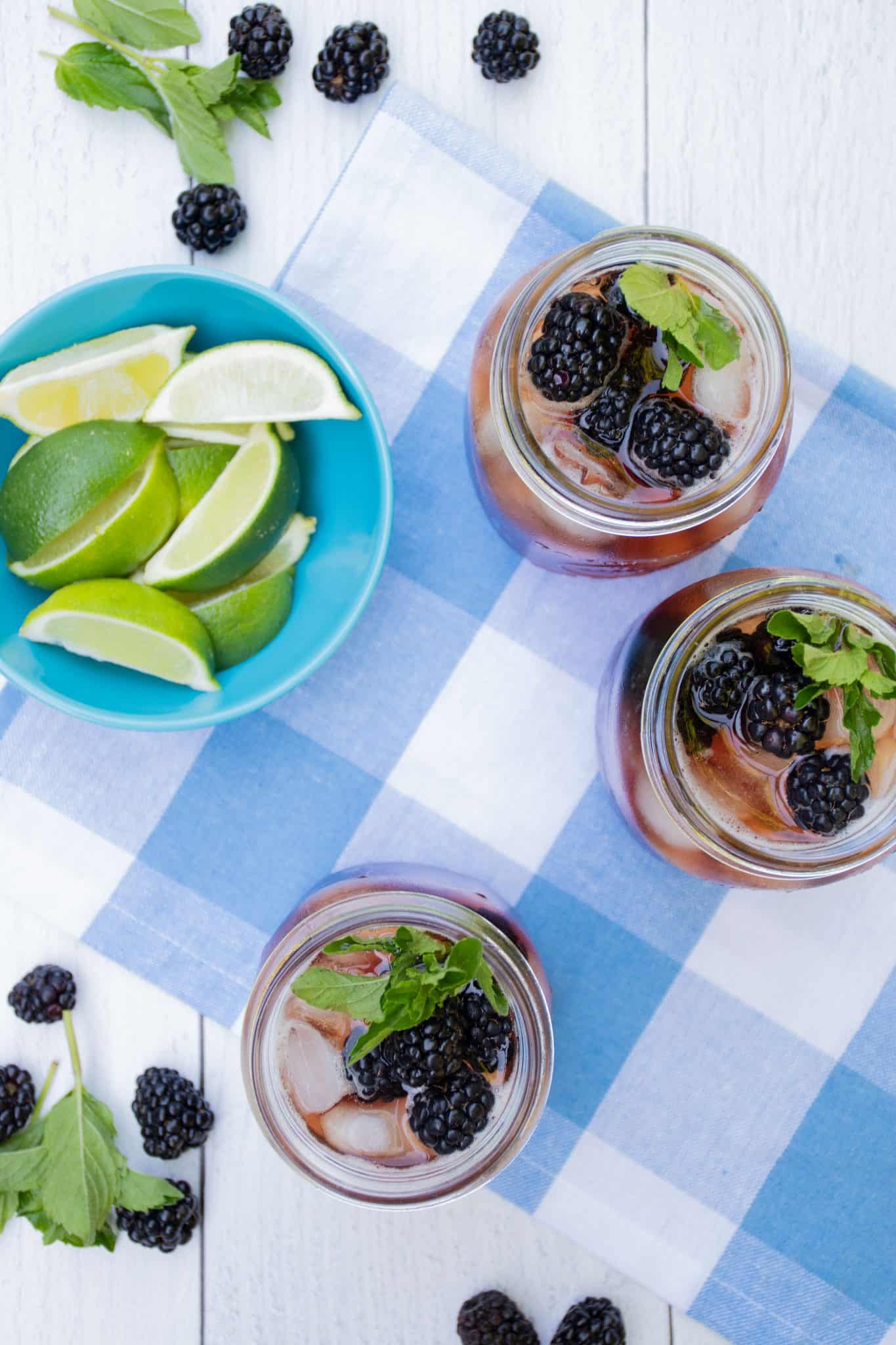 Overhead shot of iced tea in 3 Mason jars, with ice, blackberries, and mint. Small bowl of lime slices to the side.