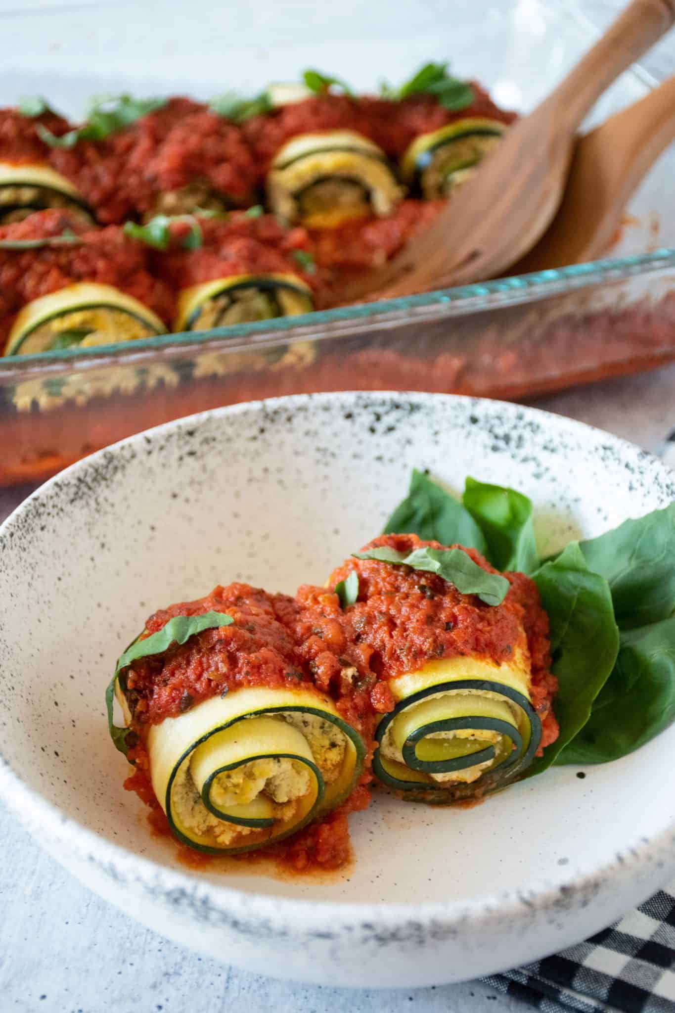 Two zucchini rolls in a bowl, with pan of rolls in background.