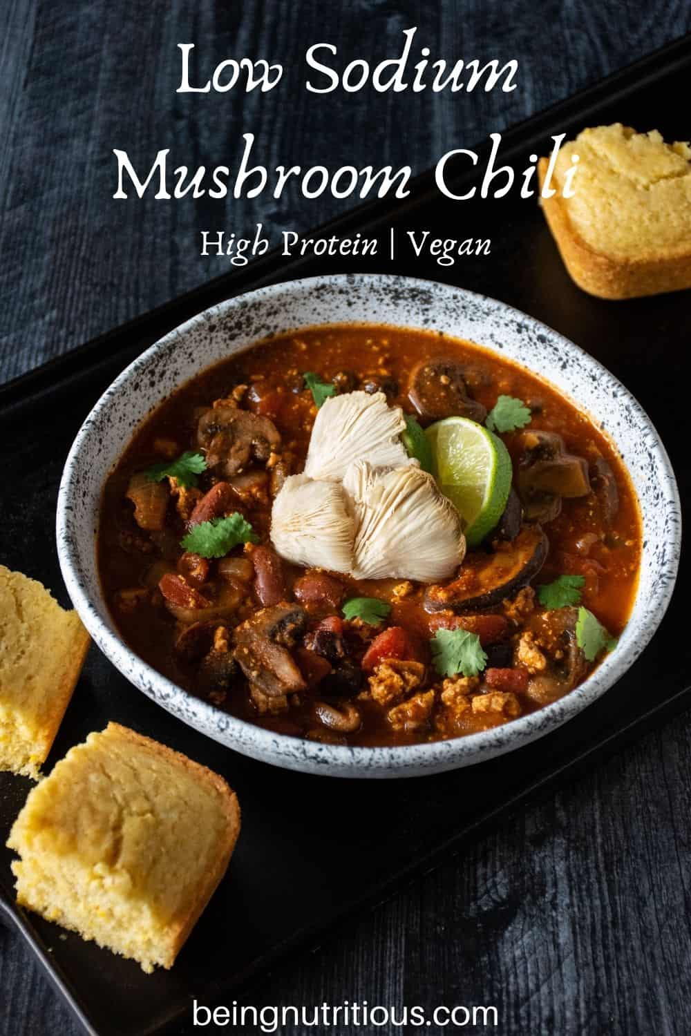 Bowl of chili with squares of cornbread surrounding. Text overlay: Low Sodium Mushroom Chili; high protein, vegan.