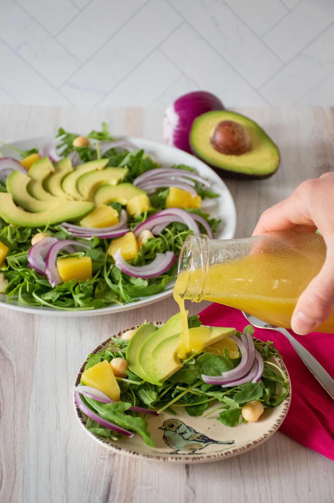 Salad on a small serving dish with dressing being poured on. Text overlay: Arugula Avocado Salad with Pineapple Vinaigrette..