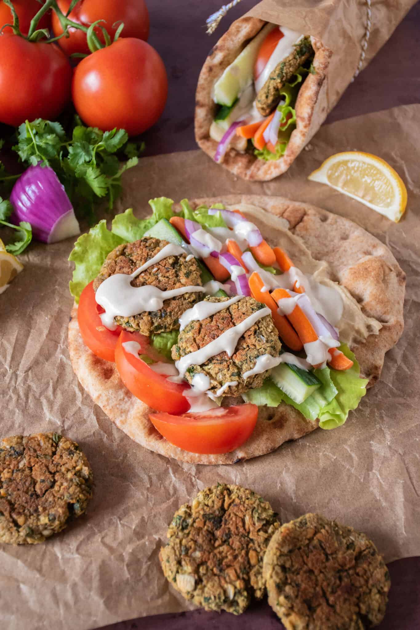 Open flatbread with lettuce, tomato, carrots, onions, cucumbers, and falafel with tahini dressing drizzled over it.