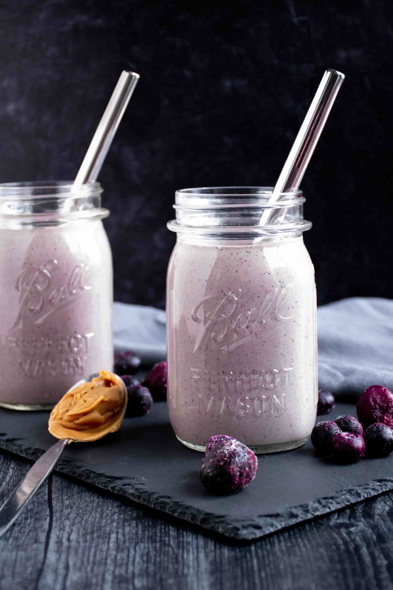 2 berry smoothies in mason jars, with frozen berries scattered around the outside, and a spoon with peanut butter on it.