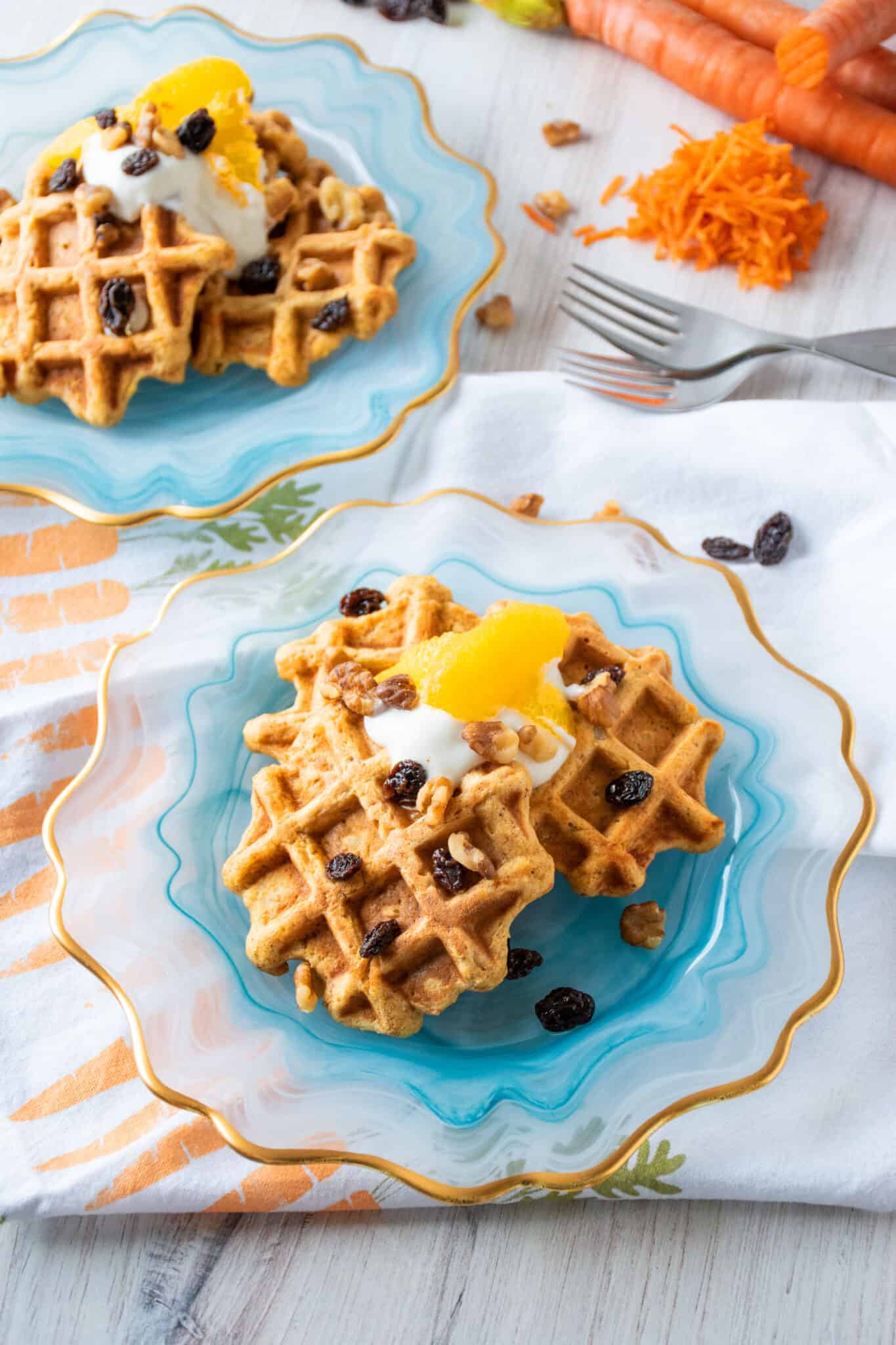 Plate of three small waffles, topped with yogurt, oranges, raisins, and walnuts. Another plate, dressed the same, and some shredded carrots are visible in the background.