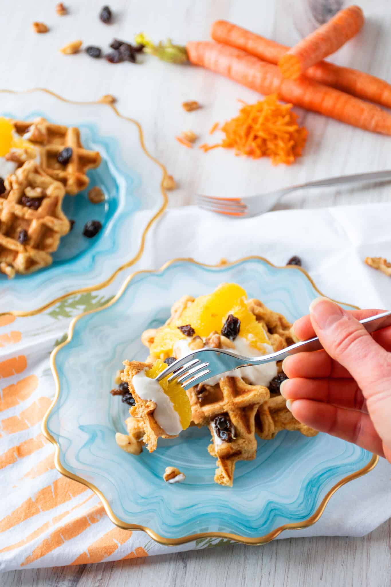 Plate of three small waffles, topped with yogurt, oranges, raisins, and walnuts. A hand is holding a fork with a waffle slice, some yogurt, and an orange slice on it in front of the plate.