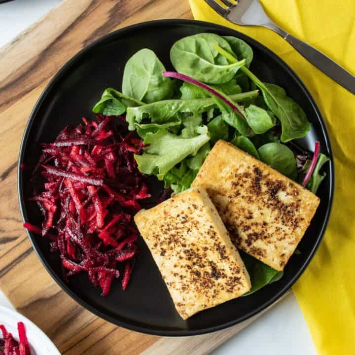 Overhead image of a plate with 2 slices of cooked tofu over a bed of greens, with a side of shredded beet salad.