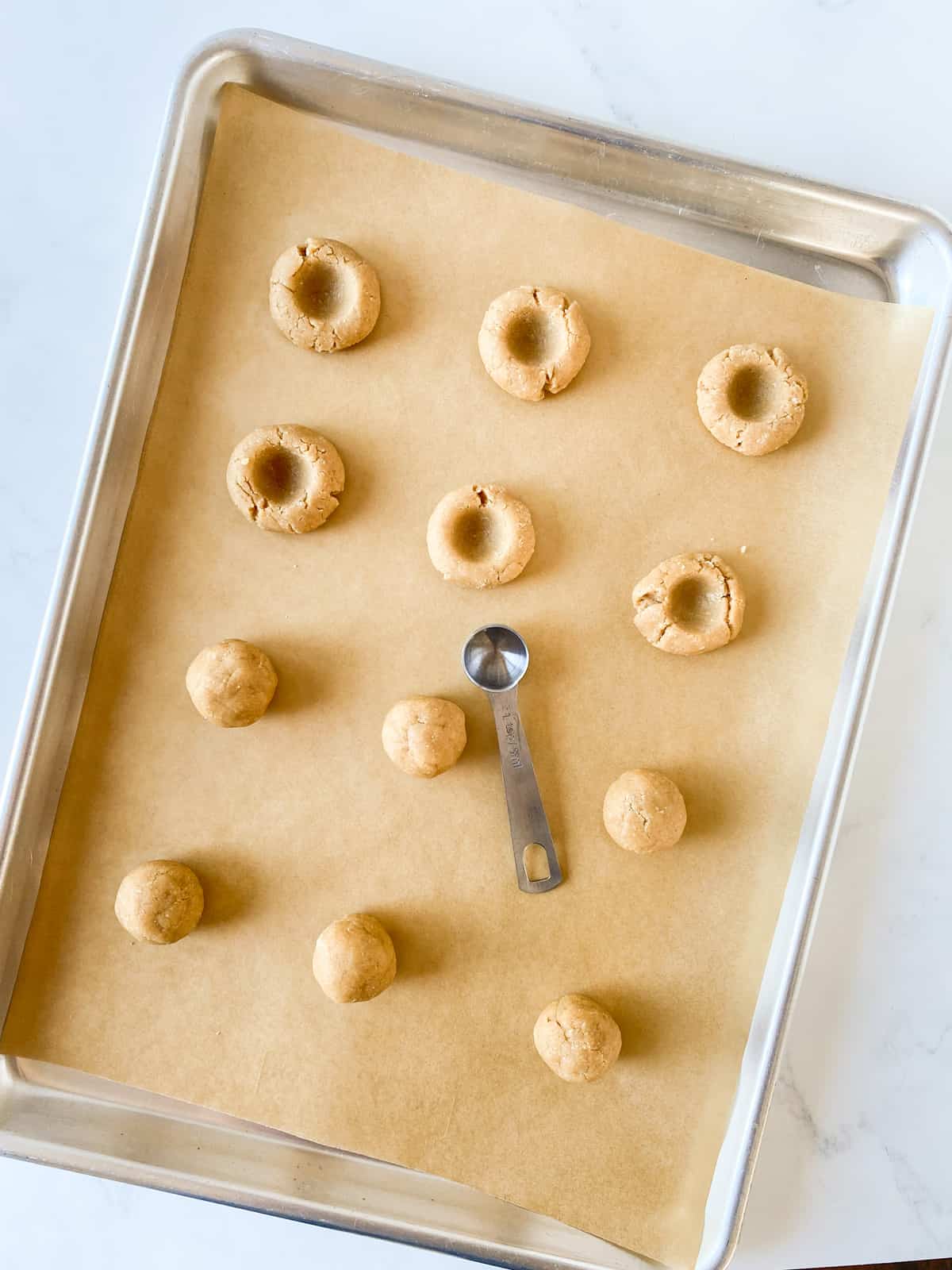 Unbaked cookies on a cookie sheet, showing using a teaspoon to make indentations in thumbprint cookies.