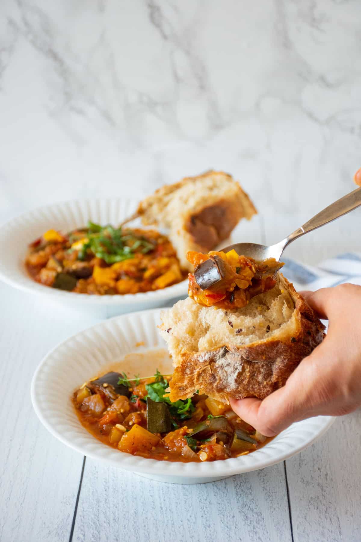 Scooping a spoonful of ratatouille onto a piece of crusty bread from a bowl.