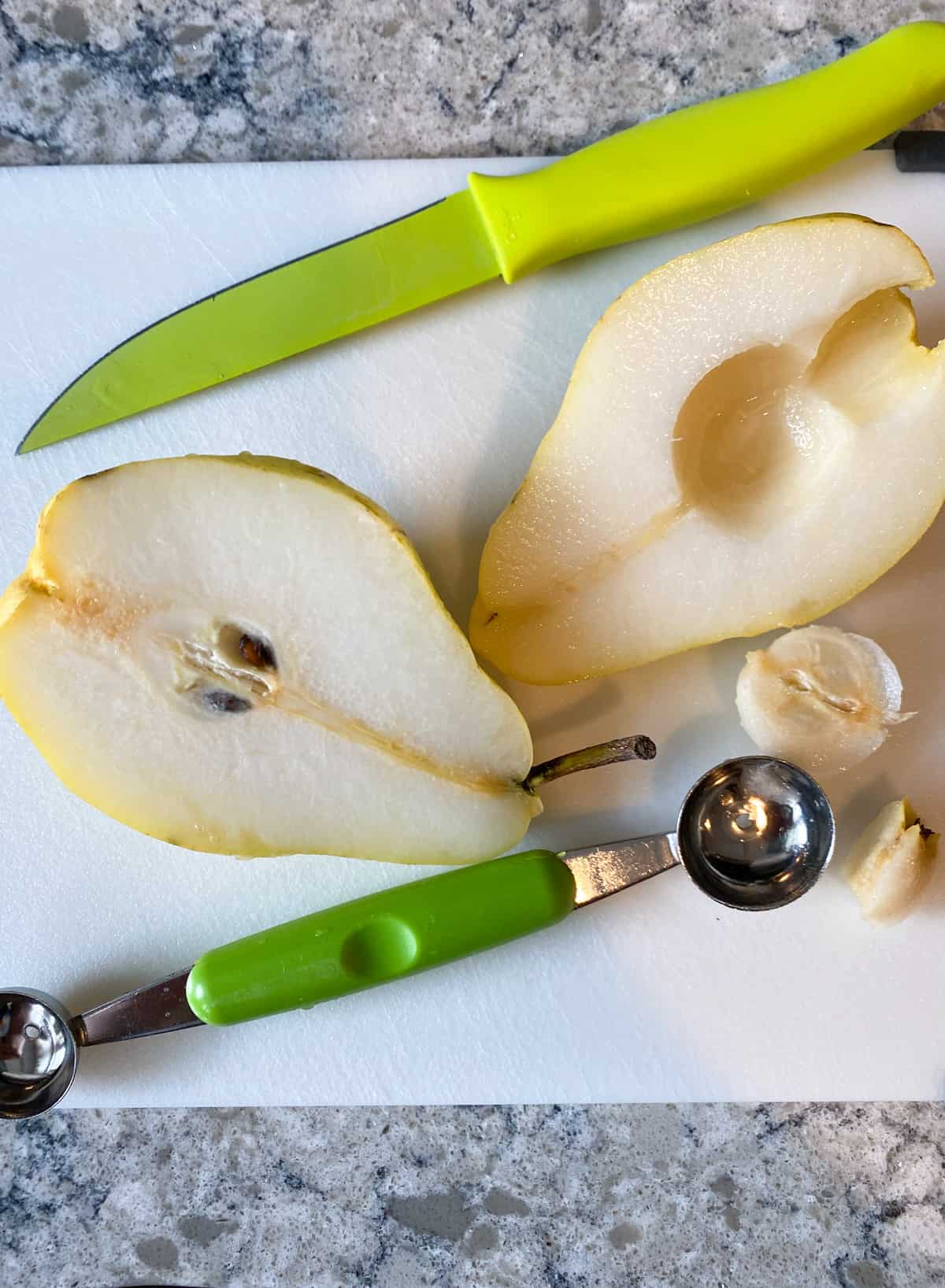 Pear cut in half on a cutting board, with a melon baller, and one half of the pear with the core removed.