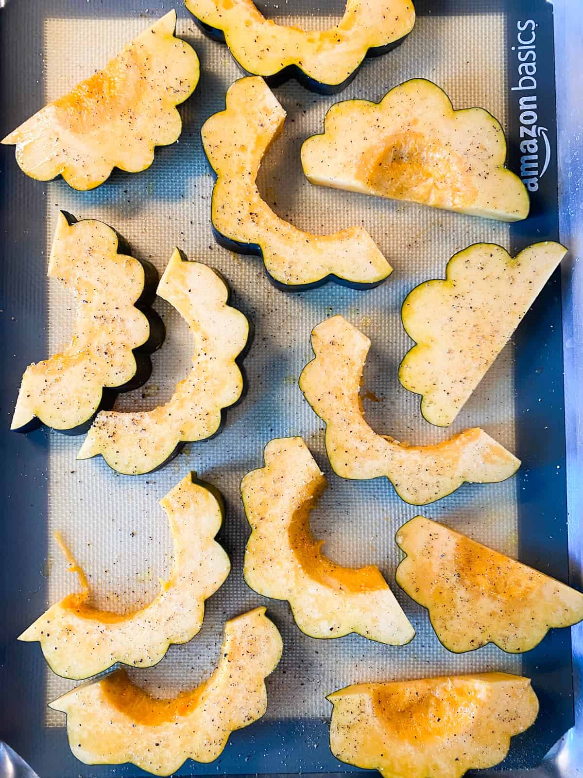 Sliced, raw acorn squash, on a baking sheet, oiled, with ground pepper.