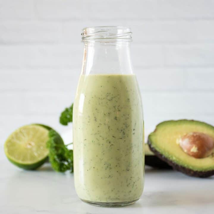 Jar of avocado salad dressing, with avocado, limes, and parsley in the background.