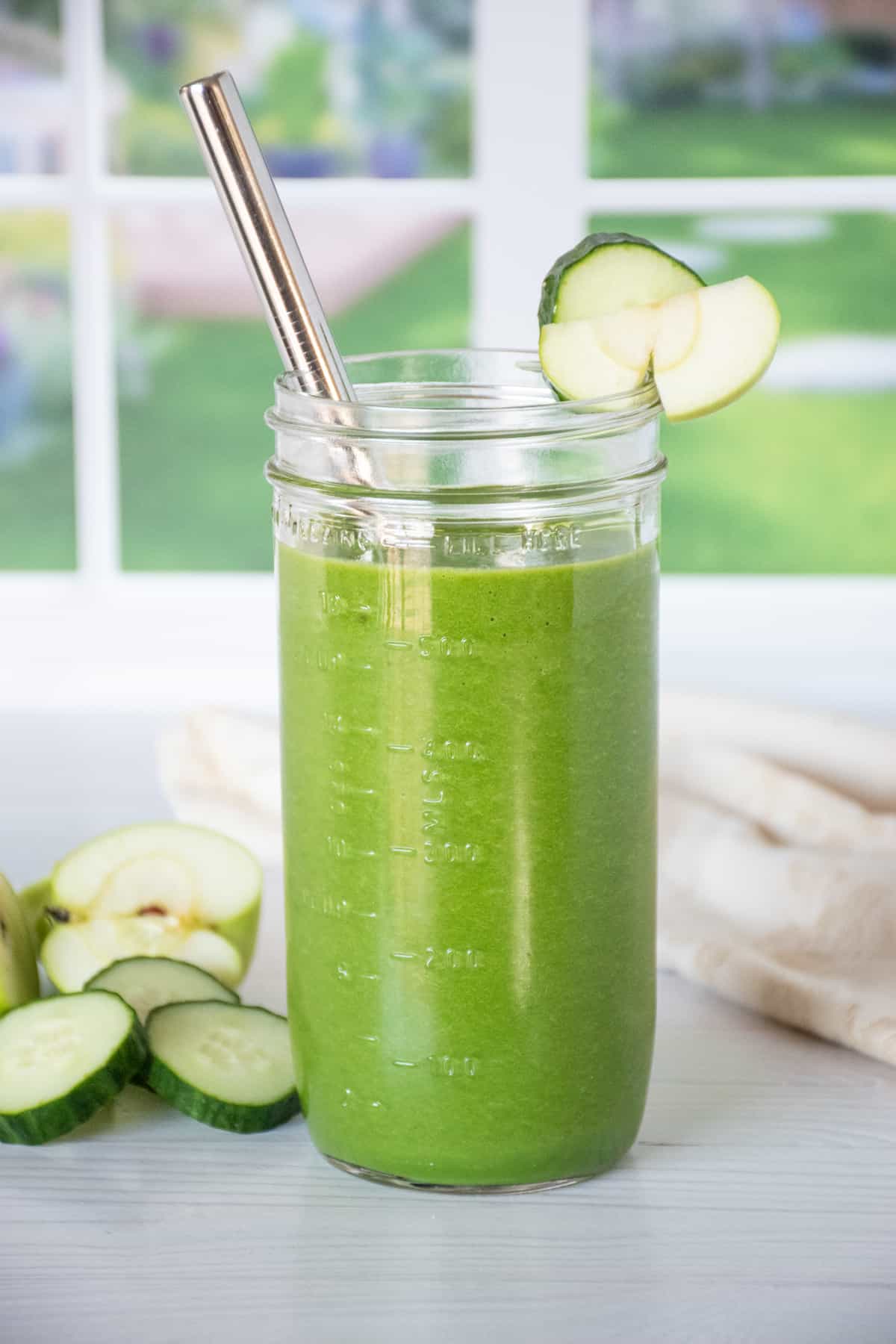 A green smoothie in a tall Mason jar with a metal straw. Cucumber slices and green apple pieces in the background.