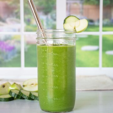 A green smoothie in a tall Mason jar with a metal straw. Cucumber slices and green apple pieces in the background.