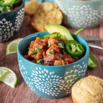 Bowl of chili, garnished with avocado, jalapeno slices, and cilantro, and a cornbread muffin on the side. Lime slices and additional bowls and spoons visible in the background.