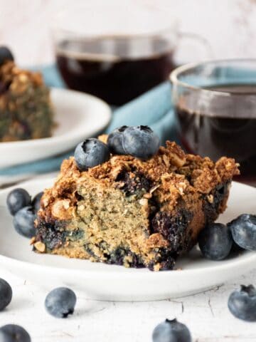 Slice of coffee cake on a plate, garnished with blueberries, with another slice of cake and two cups of coffee in the background.