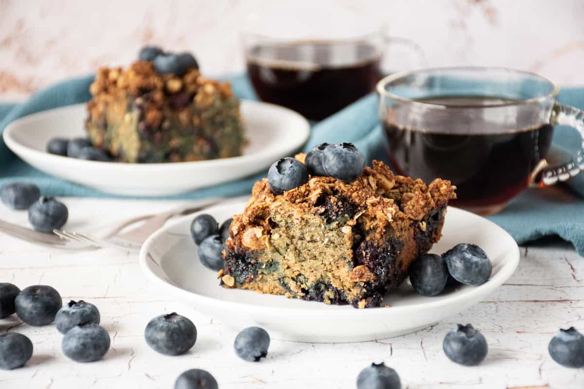 Slice of coffee cake on a plate, garnished with blueberries, with two cups of coffee and another cake slice on a plate in the background.