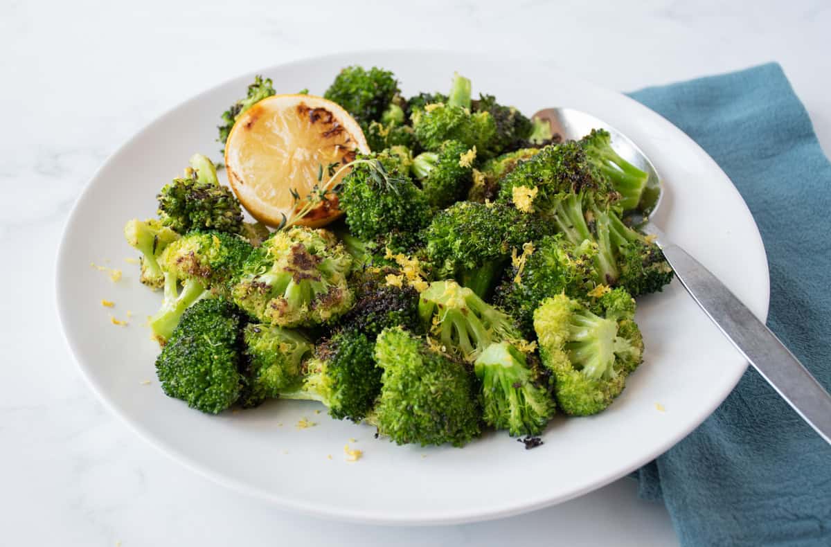 Grilled broccoli on a serving plate, garnished with a grilled lemon half, lemon zest and a stem of thyme.