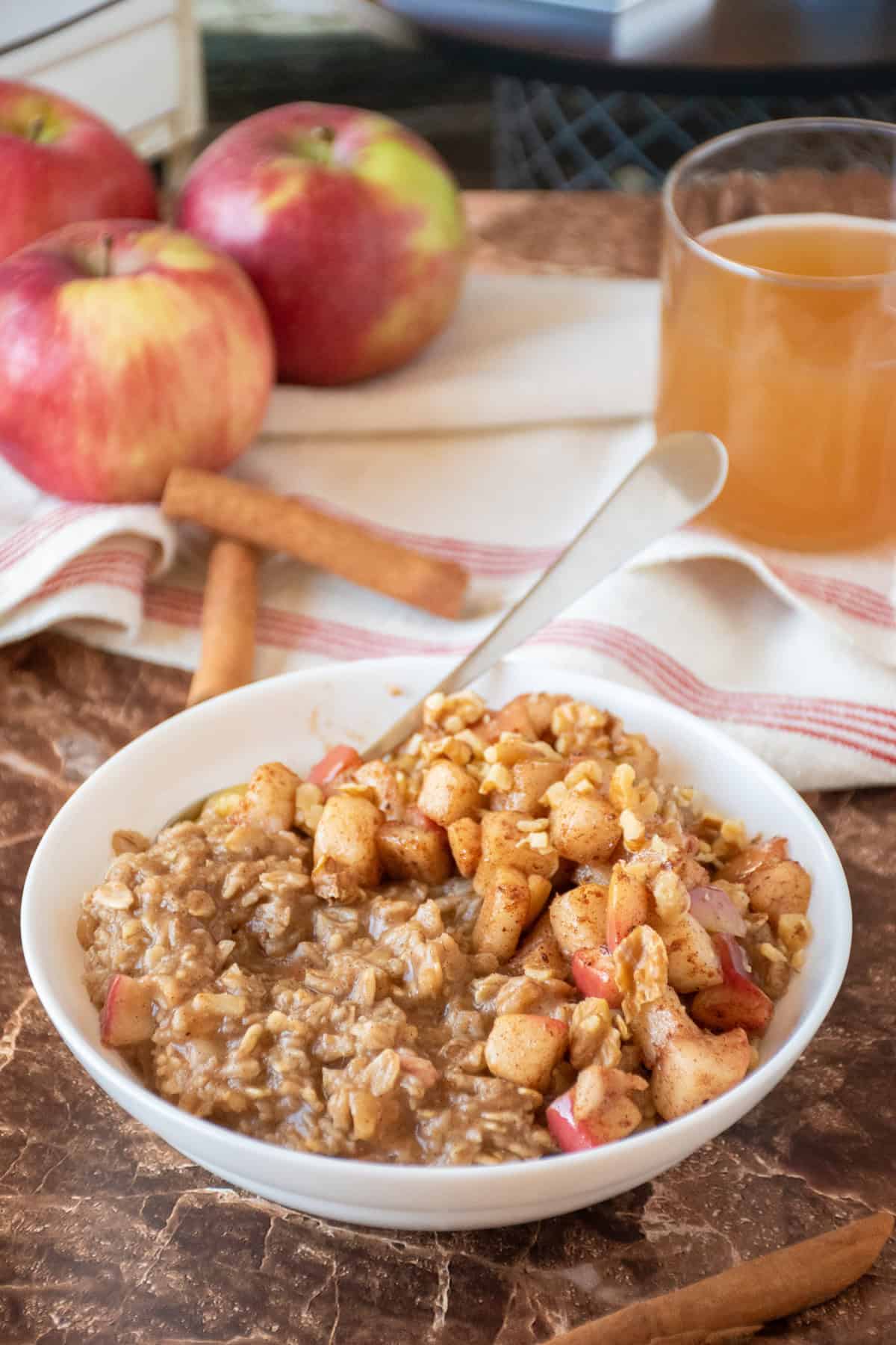 Bowl of oatmeal with sauteed apples and walnuts on top. A glass of apple cider and some whole apples in the background.