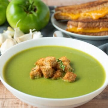 Green soup in a bowl, garnished with homemade croutons and fresh thyme. A grilled cheese sandwich and fresh garlic and green tomatoes are in the background.