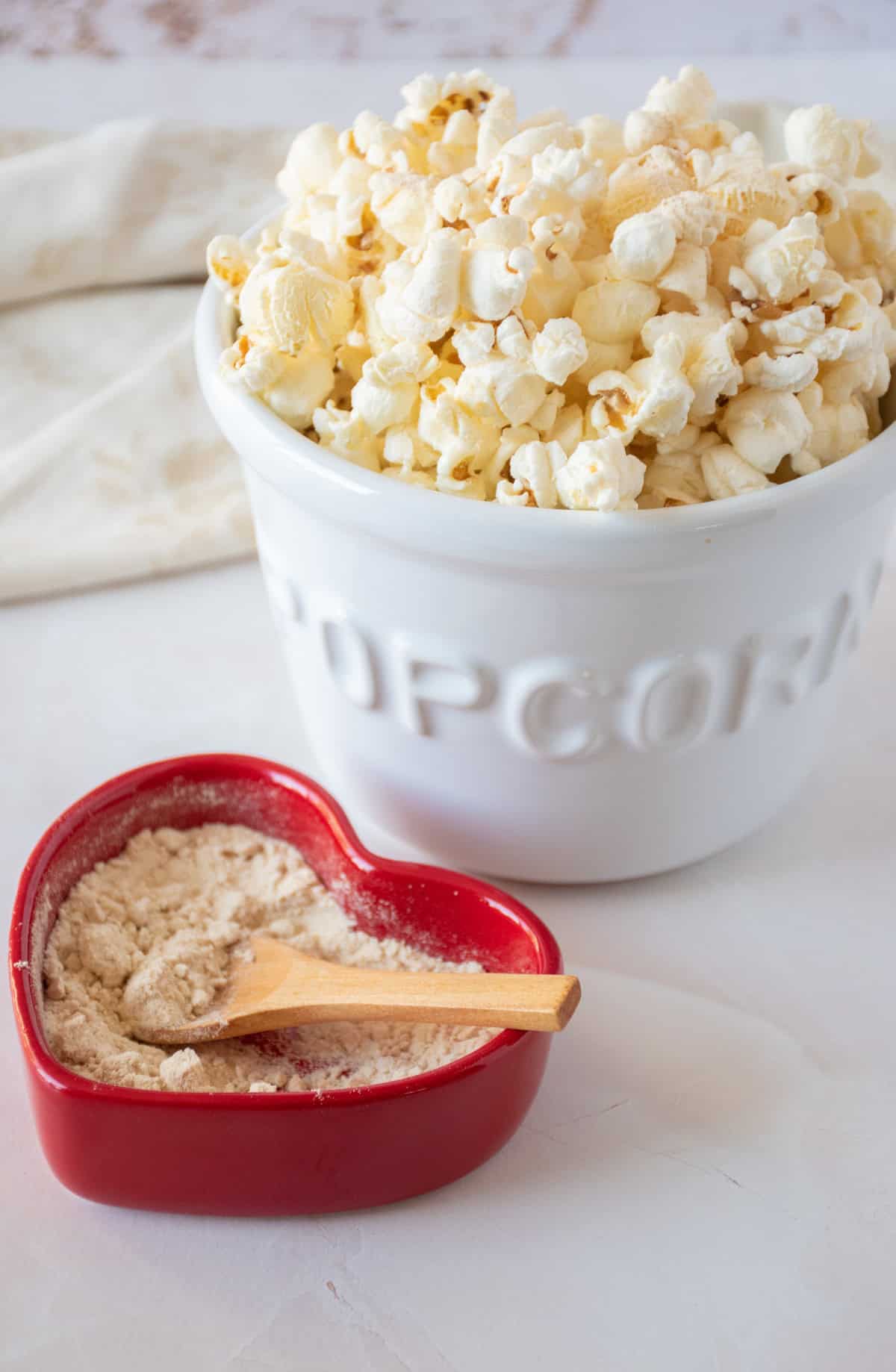 Seasoning powder made from pears and ginger, in a red heart shaped dish with a small wooden spoon. A large bowl with popcorn in it is nearby.