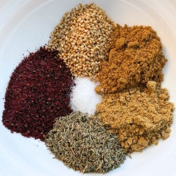 Small piles of 6 individual herbs and spices for za'atar seasoning in a white bowl.