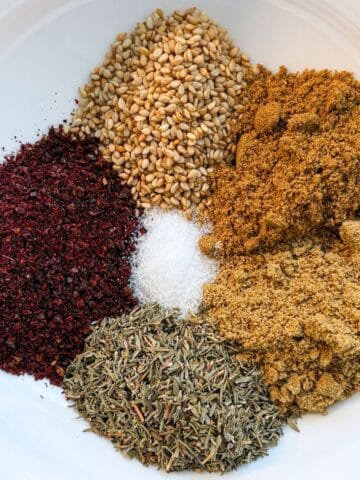 Small piles of 6 individual herbs and spices for za'atar seasoning in a white bowl.