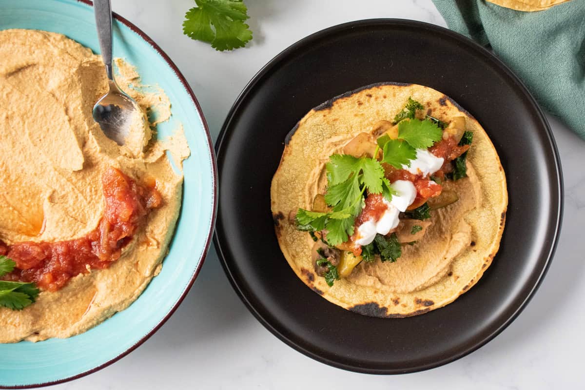 Tortilla, flat on a plate, smeared with hummus and loaded with sauteed mushrooms, zucchini, and kale, garnished with cilantro. Bowl of hummus nearby.