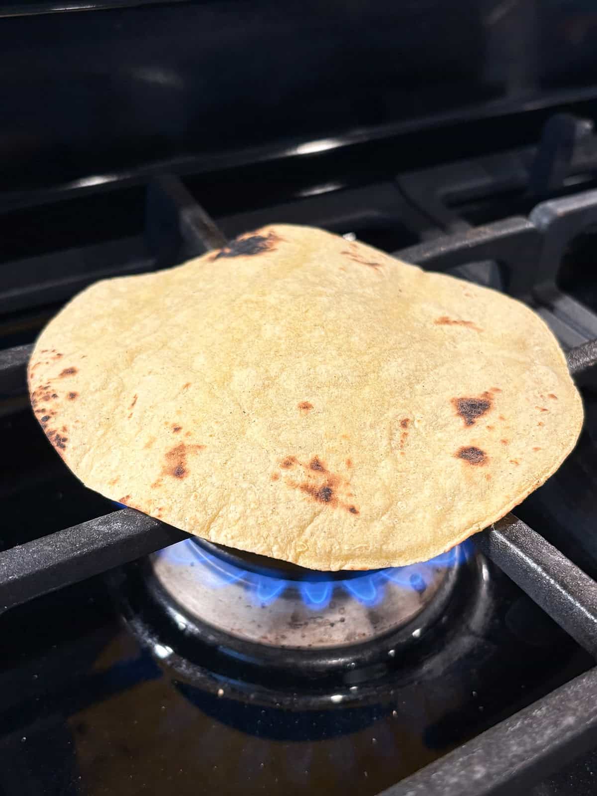 Corn tortilla on a gas stove burner, getting slightly charred.