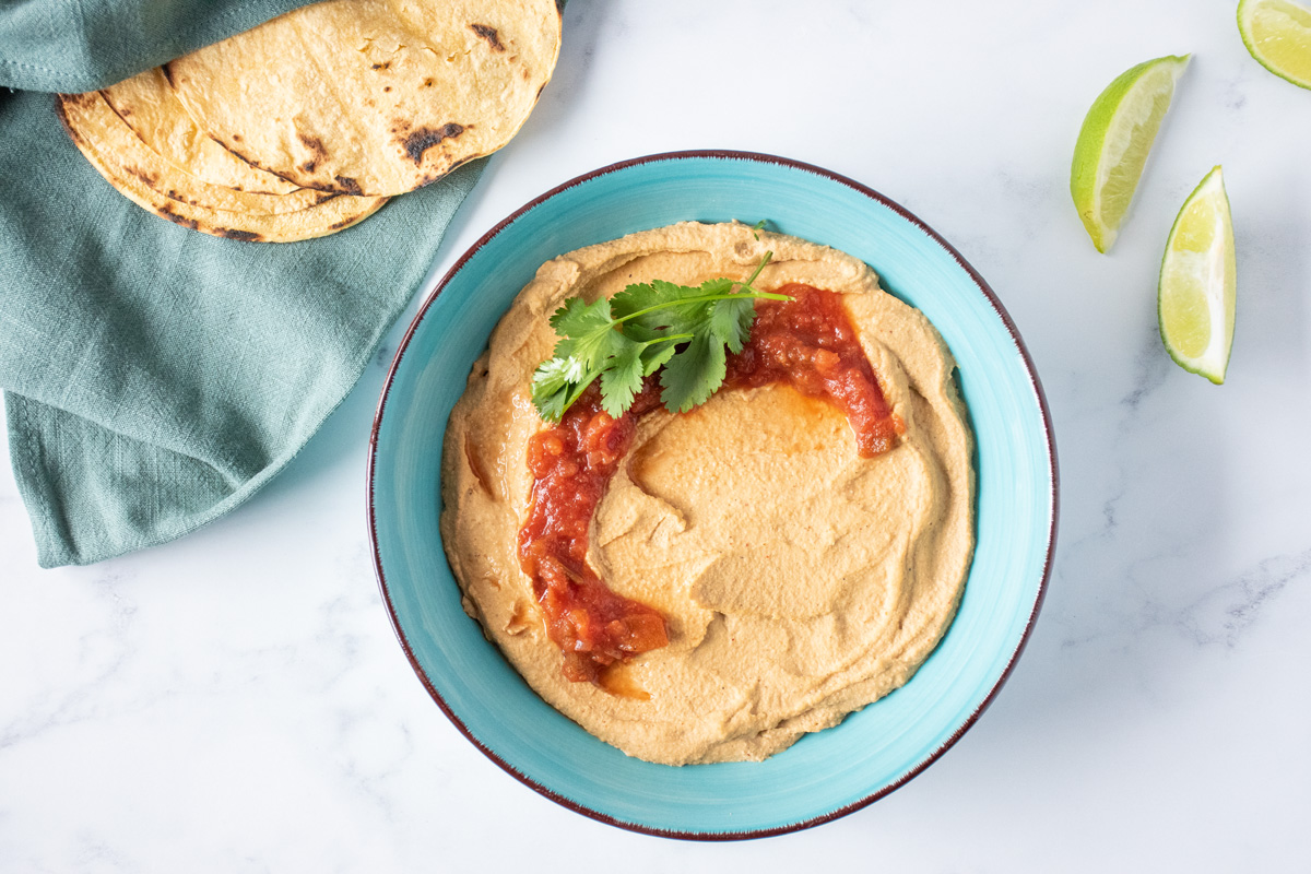 Bowl of hummus, garnished with salsa and cilantro. Cut limes and corn tortillas surround the bowl.