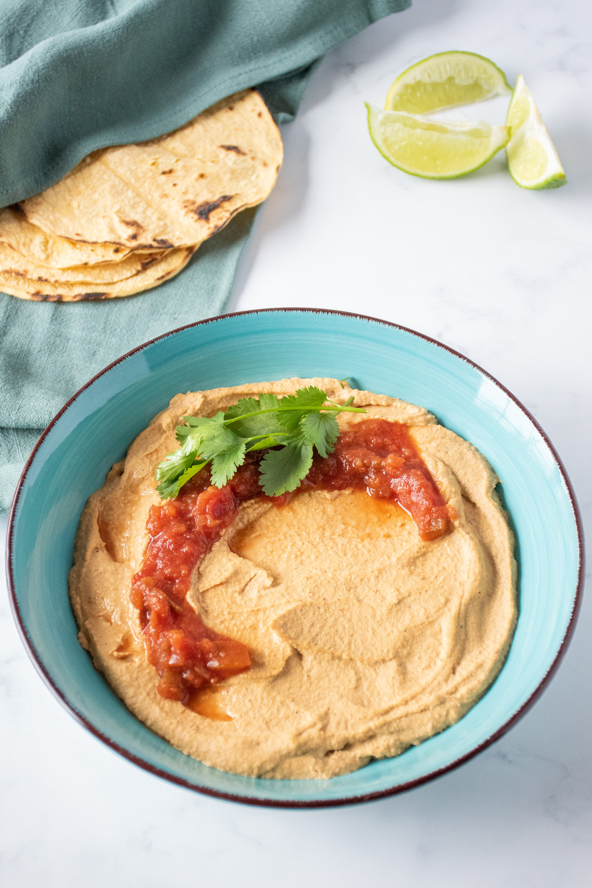 Bowl of hummus, garnished with salsa and cilantro. Cut limes and corn tortillas surround the bowl.