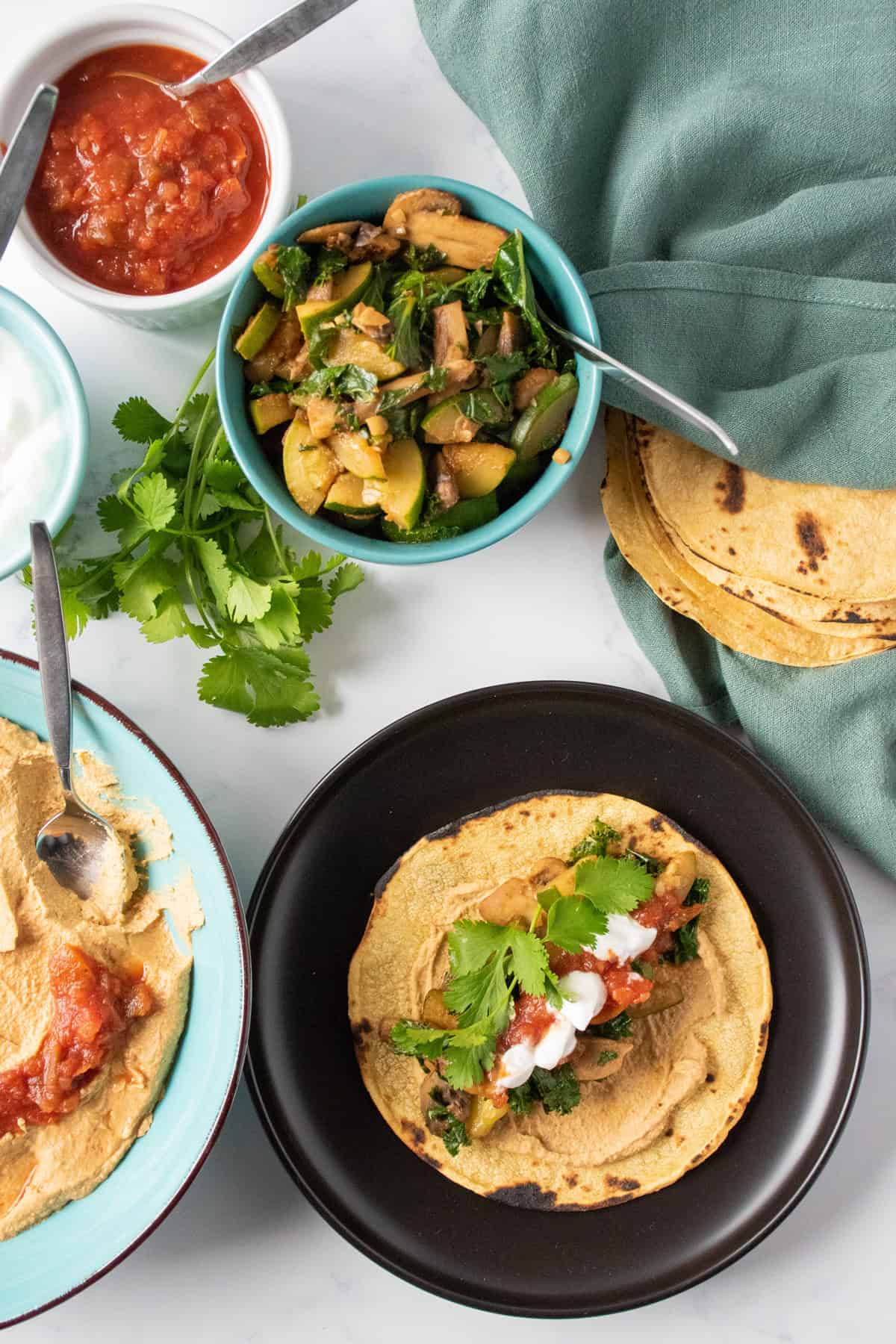 Tortilla, flat on a plate, smeared with hummus and loaded with sauteed mushrooms, zucchini, and kale, garnished with cilantro. Bowls with different fillings surround the plate.
